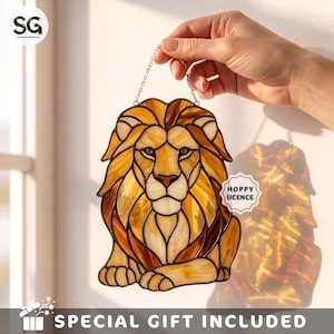 May include: A stained glass lion suncatcher featuring shades of amber, gold, and brown glass pieces joined by dark metal solder lines. The lion is depicted in a seated position and includes a small white sticker with the text HOPPY LICENCE.