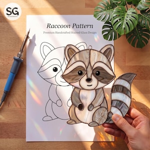 May include: A stained glass raccoon suncatcher featuring shades of beige, brown, and white glass with dark solder lines. Beside it lies a paper template titled Raccoon Pattern Premium Handcrafted Stained Glass Design and a soldering iron.