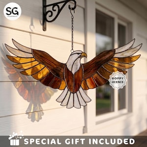 May include: A stained glass eagle suncatcher with wings spread wide, featuring white, brown, and amber glass panels held by dark metal cames. The eagle hangs from a black metal chain and wall bracket. A small white tag reads HOPPY LICENCE.
