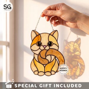 May include: A stained glass suncatcher shaped like a sitting cat with orange and cream-colored glass pieces held together by dark metal solder lines. The cat has large black eyes and a curled tail, suspended by a silver chain for hanging display.