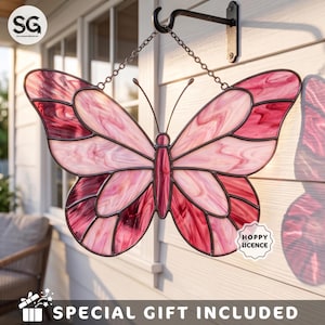 May include: A stained glass butterfly suncatcher featuring shades of pink and white with dark metal leading. The butterfly is suspended by a metal chain from a black wall-mounted hook. The design includes two antennae and a central body section.