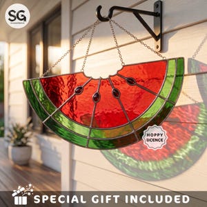 May include: A stained glass watermelon slice suncatcher featuring vibrant red and green glass segments with dark glass seeds. The piece is suspended by metal chains from a black wall-mounted bracket and includes a tag labeled HOPPY LICENCE.