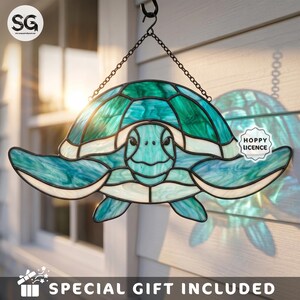 May include: A stained glass sea turtle suncatcher featuring shades of teal, turquoise, and white glass pieces held together by dark metal solder lines. The turtle is suspended by a black metal chain and hook, displaying a detailed shell pattern.