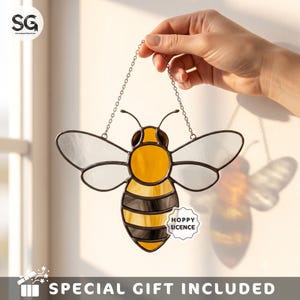 May include: A stained glass bee suncatcher featuring a yellow and amber body with black stripes and clear, textured wings. The piece is framed in dark metal solder and hangs from a silver-toned chain. A small white label reads HOPPY LICENCE.
