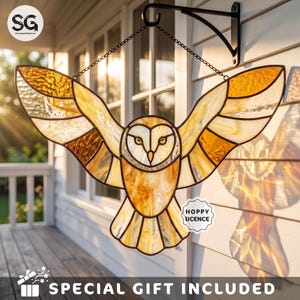 May include: A stained glass owl suncatcher with outstretched wings in shades of amber, gold, and cream. The owl features a heart-shaped face and is suspended by a black metal chain from a matching black wall-mounted bracket, casting a shadow.