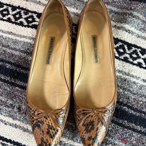 Pode incluir: Um par de sapatos rasos com padrão de pele de cobra em castanho e preto, com biqueira pontiaguda e um pequeno laço decorativo na frente. O interior dos sapatos apresenta um forro bege com o nome da marca Manolo Blahnik impresso a preto.