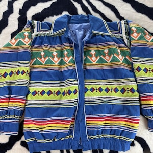 Puede incluir: Una chaqueta azul de manga larga con un vibrante patrón horizontal de formas geométricas en verde, amarillo, rojo y blanco. La prenda cuenta con un cuello clásico, cierre frontal de botones y puños y dobladillo elásticos con textura acolchada.
