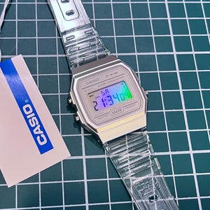 Peut inclure: Une montre numérique Casio avec un bracelet transparent et un cadran argenté. L'écran affiche l'heure en numérique avec des couleurs bleues, vertes et violettes. Une étiquette blanche avec le logo Casio est attachée.