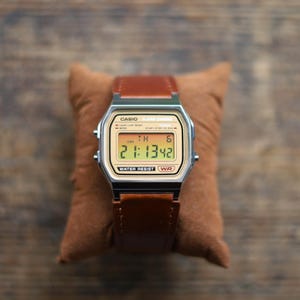 May include: A digital wristwatch with a silver-tone rectangular case and a brown leather strap. The watch face features a gold-toned background with the text CASIO, ALARM CHRONO, and WATER RESIST. The digital display shows the time 21:13:42.