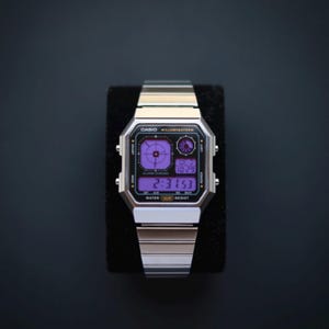 Puede incluir: Reloj digital Casio Illuminator plateado con pantalla púrpura. El reloj muestra la hora 2:31:53 y la fecha. El reloj tiene una correa de metal plateado y se muestra sobre una superficie negra.
