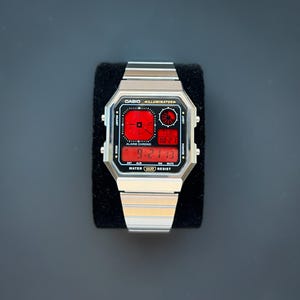 Puede incluir: Un reloj digital Casio plateado con una esfera rectangular y una pantalla digital roja. La esfera muestra la hora, la fecha y otras funciones. El reloj tiene una correa de acero inoxidable y está sobre un fondo oscuro.