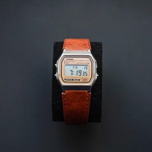 May include: A digital watch with a brown leather band. The watch face is silver with a gold-toned display showing the time and date. The watch is labeled "CASIO" and "WATER RESIST WR".