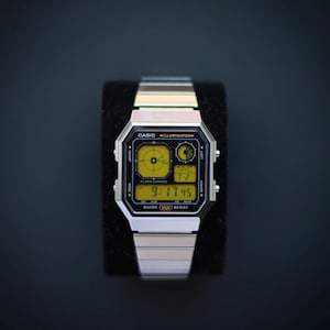 May include: A silver Casio digital watch with a black and yellow display. The watch face shows the time, 9:17:45, and other functions. The watch has a silver metal strap and is displayed on a black surface.