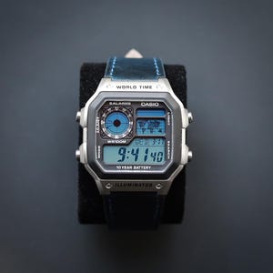 Puede incluir: Un reloj digital Casio plateado con una correa de cuero azul. La esfera del reloj muestra la hora, la fecha y las funciones de hora mundial. El reloj tiene una esfera rectangular con botones laterales y el texto "WORLD TIME" y "CASIO".