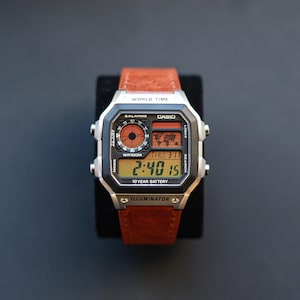 May include: A digital Casio watch with a brown leather strap. The watch face shows the time, date, and world time features. The watch has a silver-tone case and a black display with orange accents.