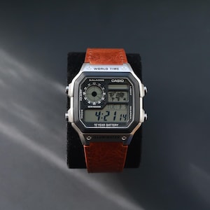 May include: A Casio digital watch with a brown leather strap. The watch face displays the time, date, and world time features. The watch has a silver-tone case and black digital display. The watch is on a black surface.