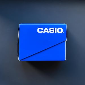 以下が含まれることがあります： 鮮やかな青色のCasioの箱。ブランド名は白で、箱の上部に配置されています。対角線デザインがあり、濃い青色の背景に配置されています。時計やその他の小型電子機器用である可能性が高いです。