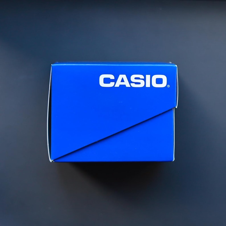 以下が含まれることがあります： 鮮やかな青色のCasioの箱。ブランド名は白で、箱の上部に配置されています。対角線デザインがあり、濃い青色の背景に配置されています。時計やその他の小型電子機器用である可能性が高いです。