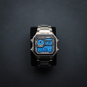 May include: A silver Casio digital watch with a black face and blue digital display. The watch shows the time as 13:06:17. The watch has a stainless steel strap and is displayed on a black surface.
