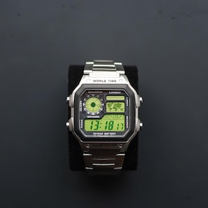 Könnte beinhalten: Eine silberne Casio Digitaluhr mit einem schwarz-grünen Display, das die Uhrzeit, das Datum und eine Weltkarte anzeigt. Die Uhr hat ein Edelstahlarmband und ein schwarzes Zifferblatt mit dem Text "CASIO" und "WORLD TIME".