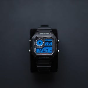 May include: Black Casio digital watch with a blue digital display. The watch features multiple functions, including world time and a light. The display shows the time as 12:00-4. The watch is water resistant to 100 metres.