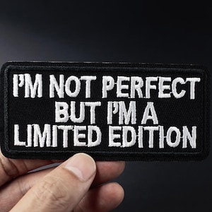 Peut inclure: Écusson rectangulaire noir avec texte brodé blanc : "I'M NOT PERFECT BUT I'M A LIMITED EDITION". Le patch est tenu par une main sur fond sombre. Un accessoire de mode pour vêtements.