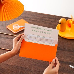 May include: A bright orange envelope containing a white card with the text 5 Daily Habits to Reduce Stiffness at Your Desk. The card includes a bullet point titled Move Every Hour with instructions to take short breaks to stretch or walk.