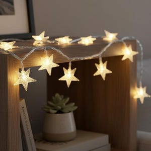 Könnte beinhalten: Eine Lichterkette mit warmweißen sternförmigen Lichtern liegt über einem Nachttisch aus Holz. Die Lichter sind eingeschaltet und erzeugen ein sanftes Leuchten. Ein kleiner Topf mit Sukkulenten und Bücher befinden sich ebenfalls auf dem Tisch.