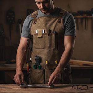 May include: A brown canvas apron with multiple pockets, leather straps, and a leather neck strap. The apron is worn by a person in a workshop. Tools, including pliers, wrenches, and pens, are visible in the pockets. A large wrench is on the wooden surface.