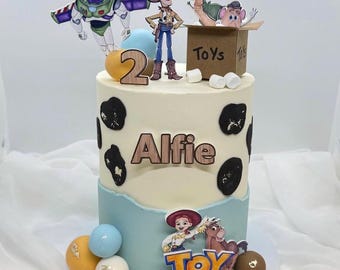 Può includere: Una torta di compleanno a due piani decorata con personaggi di Toy Story. La torta ha una base azzurra e una parte superiore bianca con macchie nere. La torta è decorata con il nome "Alfie" e il numero "2".