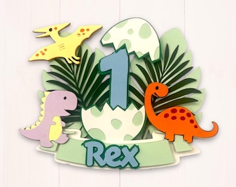 May include: A colorful dinosaur-themed cake topper featuring a number 1, an egg, and three dinosaurs. The topper includes the name "Rex" on a banner, with a yellow Pterodactyl, a purple dinosaur, and an orange dinosaur.