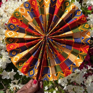 Pode incluir: Um leque de mão colorido com um design geométrico em vermelho, amarelo, azul e verde. O leque é exibido em um fundo de flores brancas e vermelhas, criando um contraste vibrante. O design do leque é intrincado e atraente.
