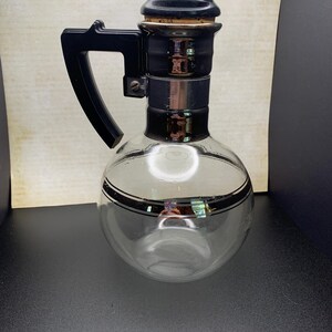May include: A vintage clear glass coffee carafe with a black handle, lid, and accents. The carafe has a round base and a black band around the middle. The lid is a dark, rounded shape.