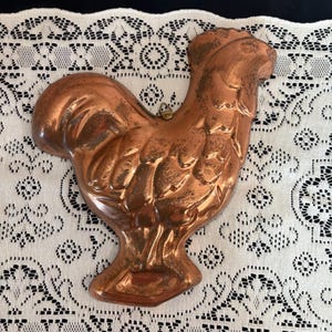 May include: A copper-colored rooster-shaped mold. The mold has detailed feather and body features. It is hanging on a white lace tablecloth. The rooster mold is a vintage kitchenware item.