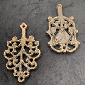 May include: Two antique cast iron trivets. One is a decorative openwork design, the other is shield-shaped with a spade and utensil motif. Both have a rustic, aged appearance with a bronze-colored finish.