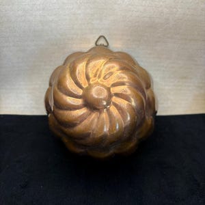 May include: A copper-colored, antique bundt pan with a decorative, swirled design. The pan has a central circular indentation and a small loop for hanging. The pan is displayed against a black surface and a light-colored background.