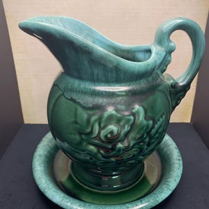 May include: A vintage green ceramic pitcher and matching saucer set. The pitcher has a rounded body, a curved spout, and a decorative handle. The set is glazed in shades of green, with darker accents. The saucer is round and complements the pitcher's design.
