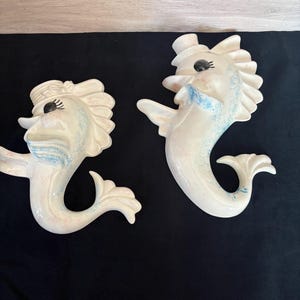 May include: Two ceramic fish wall hangings. One fish wears a top hat, the other a bow. Both are off-white with blue and pink accents. They have black eyes and are smiling. The fish have a whimsical, vintage aesthetic.