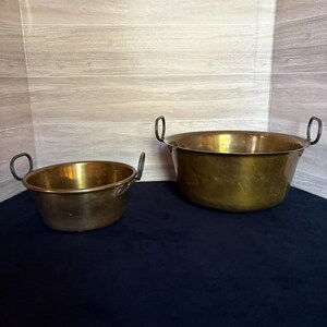 May include: Two antique brass bowls with curved handles. The bowls have a rounded shape and a shiny, golden finish. The smaller bowl is on the left, and the larger bowl is on the right. The bowls are sitting on a dark blue surface.