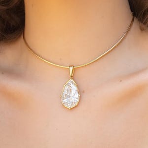 May include: A gold necklace featuring a teardrop-shaped pendant with a large, clear, faceted gemstone. The pendant is set in a gold frame. The necklace has a delicate, elegant design.