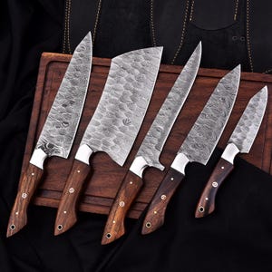 May include: A set of six kitchen knives with patterned steel blades and wooden handles. The knives are arranged on a wooden cutting board, showcasing their various sizes and shapes. The handles are a dark brown color.