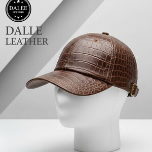 May include: A brown leather baseball cap with a crocodile skin texture. The cap has a curved brim and a brass-coloured buckle on the back. The text "DALLE LEATHER" is printed on the side.