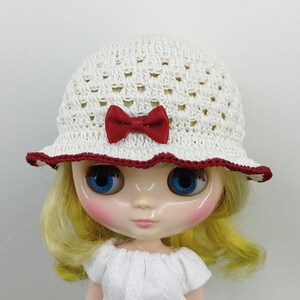 May include: A doll with blonde hair wearing a white crocheted bucket hat. The hat features an open-weave pattern, a small red satin bow centered on the front, and a dark red crocheted trim along the edge of the brim. The doll wears a white top.