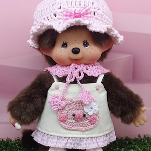 以下が含まれることがあります： ピンク色のリボン付きかぎ針編みの帽子、ピンク色の襟、豚のデザインの白いエプロンドレスを着たぬいぐるみ。人形は茶色の毛皮、笑顔の顔、白い靴をしています。背景はピンクと緑です。