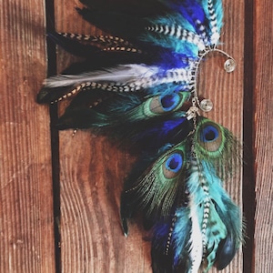 Puede incluir: Un brazalete de oreja artesanal con una vibrante variedad de plumas en tonos azules, turquesas, negros y blancos. El dise&ntilde;o incorpora plumas de pavo real y un delicado marco de alambre plateado.