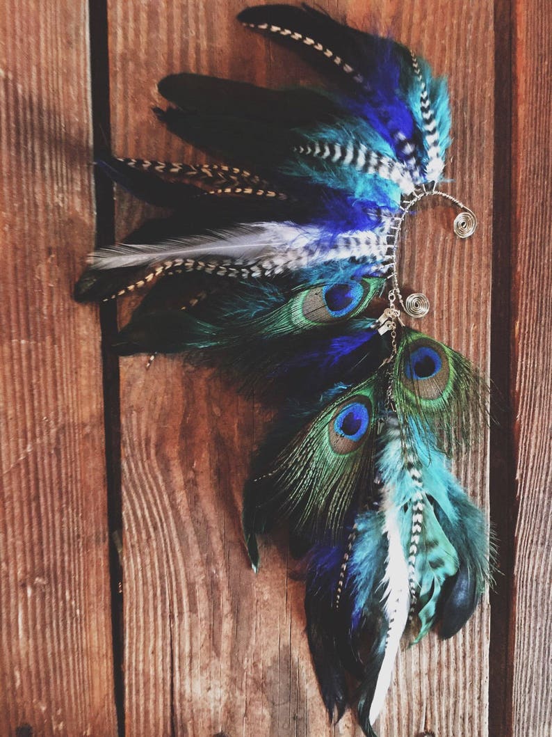 Puede incluir: Un brazalete de oreja artesanal con una vibrante variedad de plumas en tonos azules, turquesas, negros y blancos. El dise&ntilde;o incorpora plumas de pavo real y un delicado marco de alambre plateado.