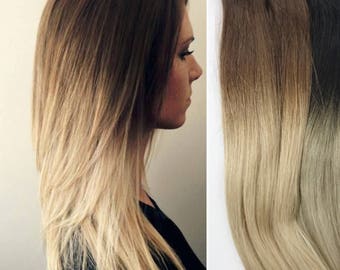 Pode incluir: Um conjunto de quatro tramas de extensões de cabelo ombré. As tramas são de cor castanho escuro nas raízes e clareiam gradualmente até um tom loiro claro nas pontas.