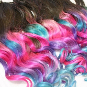 K&ouml;nnte beinhalten: Mehrere Str&auml;hnen welliger Haarverl&auml;ngerungen mit dunkelbraunen Wurzeln und Ombre-Spitzen in den Farben Pink, Blau und Lila.