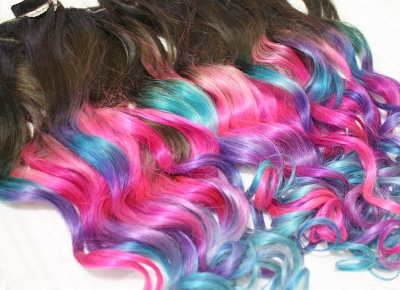 K&ouml;nnte beinhalten: Mehrere Str&auml;hnen welliger Haarverl&auml;ngerungen mit dunkelbraunen Wurzeln und Ombre-Spitzen in den Farben Pink, Blau und Lila.