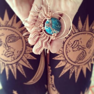 May include: A copper wire sunburst pendant with a turquoise and brown jasper stone in the center. The pendant is being held by a hand against a black background with a repeating sun design.